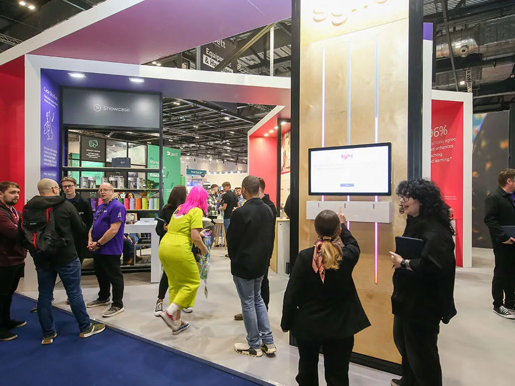 A crowd of people around a modern, angular exhibition stand. Two people stand in front of a TV screen that is a coin slot engagement