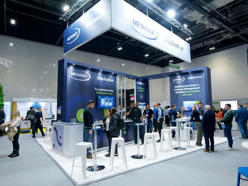 A custom exhibition stand for Metapack ShipStation at the TFM Event, featuring navy branded stand design, digital displays, and seating areas.